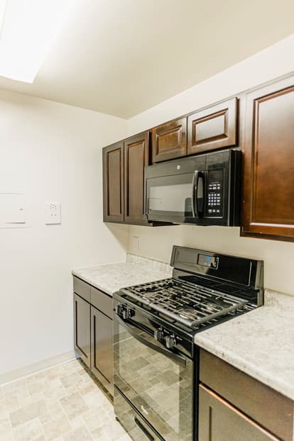 Kitchen with walnut cabinets, white walls, black appliances, tan and white floor. 